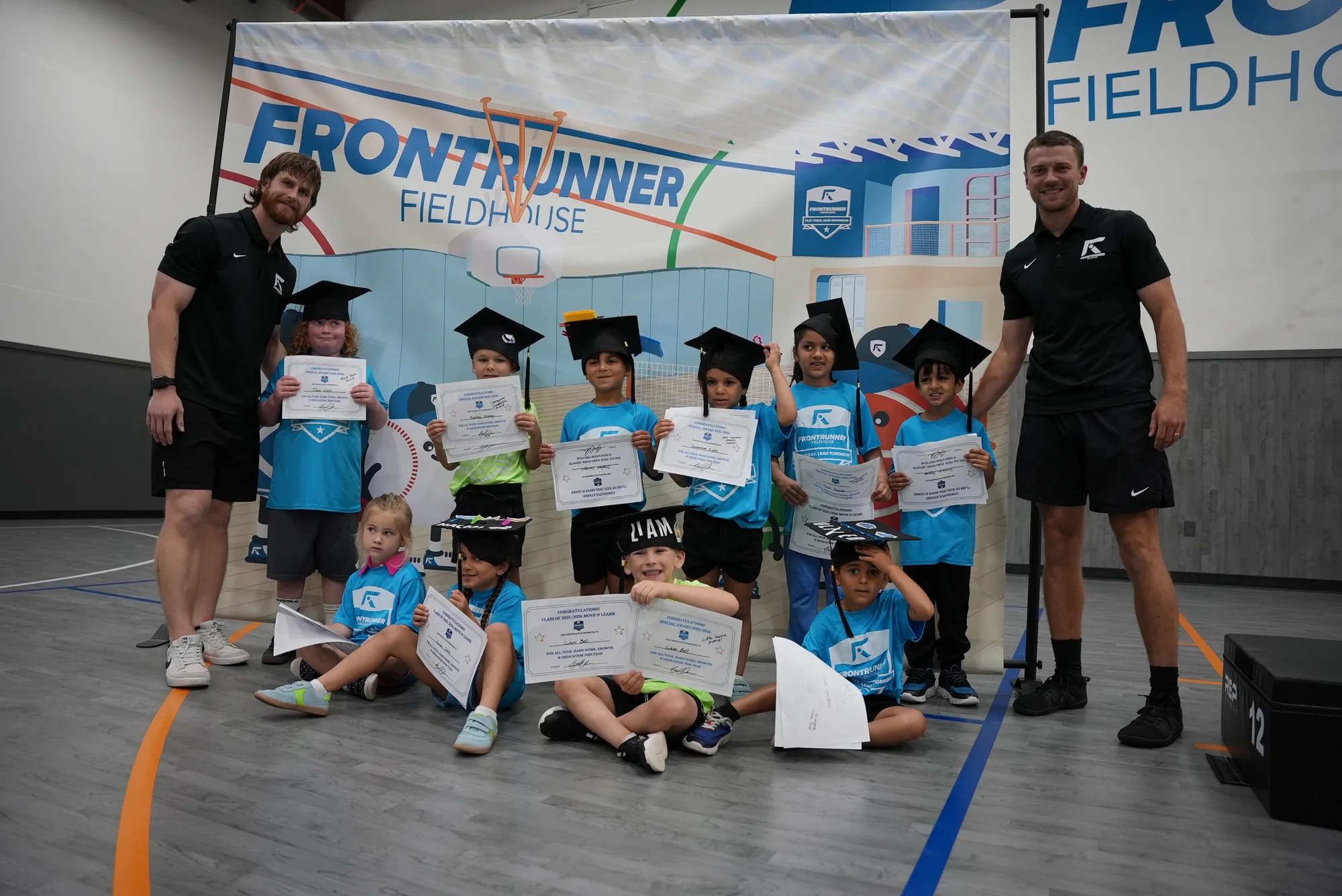 Move & Learn graduates celebrating in graduation caps under the Frontrunner Fieldhouse banner